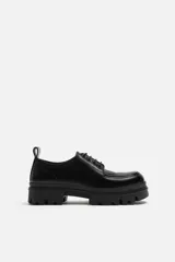 Zapato de vestir tipo blucher color negro, con detalle de bordón, tirador trasero y horma redonda. Suela con volumen y diseño irregular.