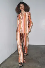 Pantalón de tiro medio con estampado de rayas verticales en tonos marrón, naranja, rosa y beige. Cuenta con trabillas en la cintura, cinturón ajustable con lazadas, bolsillos laterales y cierre frontal con cremallera y botones.