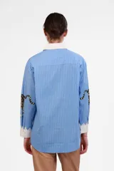 Camisa celeste a rayas verticales blancas, de corte holgado, con cuello clásico blanco, cierre frontal con botones y estampado de leopardo en los puños.