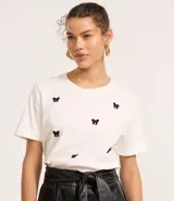 Remera baby look blanca de algodón con cuello redondo y mangas cortas. Presenta bordados de lacitos negros en el frente.