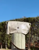 Bandolera tipo sobre o clutch, color beige metalizado o champagne, con textura brillante/iridiscente. Posee un bolsillo frontal con cierre de broche giratorio y un detalle de borla colgante en uno de los laterales. La correa es de cuero marrón oscuro.