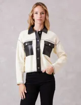 Chaqueta de corte boxy en color crema, confeccionada en tejido de punto suave con detalles en cuero sintético negro en cuello, solapas, cartera frontal y bolsillos. Presenta cierre frontal con botones metálicos plateados, hombros caídos y costuras decorativas horizontales.