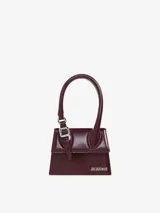 Bolso estructurado de piel lisa color borgoña, con asa reforzada de hebilla metálica, solapa magnética y asa bandolera ajustable y extraíble. Cierre de mosquetón grabado, bolsillo interior y logotipo frontal. Forrado completamente en algodón.