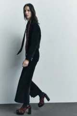 Pantalón ancho de pana color negro, de tiro medio, con trabillas en la cintura, bolsillos delanteros y cierre frontal con cremallera, botón y gancho metálico.
