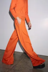 Pantalón ancho color naranja, de tiro medio y cintura con trabillas. Tiene bolsillos laterales y de vivo en espalda, pinzas en delantero, raya planchada y cierre frontal con cremallera, botón y ganchos metálicos.