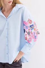 Camisa blanca de algodón con bordados florales y lentejuelas en las mangas, cuello camisero, mangas largas con puños abotonados y corte amplio.