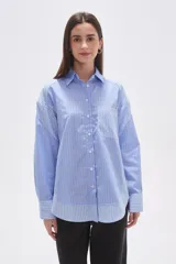 Camisa oversize de poliéster, con cuello camisero y cierre frontal de botones. Presenta un diseño de rayas verticales azules y blancas, con bloques de rayas horizontales en los puños y el ruedo inferior.