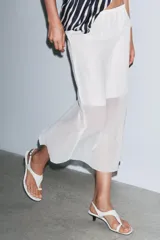 Falda midi color blanco, semitransparente, de tiro medio con cintura elástica, forro corto interior y abertura en la espalda.