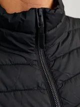 Chaleco acolchado negro sin mangas con cuello alto y cierre lateral oculto y cremallera frontal. Bolsillos frontales y diseño plegable para fácil almacenamiento. Relleno de fibra sintética para calidez ligera.