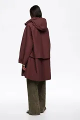 Gabardina midi de corte línea A en color bordó, confeccionada en mezcla de algodón técnico. Presenta cierre frontal con cremallera metálica, cuello subido con capucha integrada y mangas largas ajustables con botones de presión en los puños. Incluye bolsillos laterales y aberturas con botones en los costados.