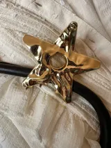 Pinza para el pelo dorada con forma de estrella. Tiene un agujero circular en el centro y una textura irregular.