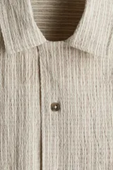 Camisa de manga corta color beige claro, confeccionada en tejido liviano de lino y algodón. Tiene cuello resort, tapeta lisa, bolsillo superior y bajo recto con aberturas laterales.