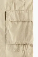Pantalón cargo color beige de lona de algodón, de tiro bajo con elástico en la parte posterior de la pretina y cierre con botón a presión. Piernas amplias con bolsillos al bies y bolsillos de fuelle con solapa.