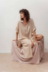 Vestido camisero largo color beige claro, de manga corta con puños doblados y abotonadura frontal. Presenta un corte holgado y aberturas laterales en el ruedo.
