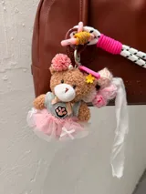 Charm de peluche con forma de oso marrón, vestido con un tutú rosa y un top gris. Lleva un detalle de huella de pata bordada en el pecho y un adorno floral en la cabeza.
