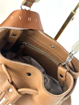 Bolso tipo bucket de cuero color marrón con cierre de cordón ajustable. Incluye dos correas intercambiables: una corta de cuero para llevar al hombro y una larga de tela con letras para usar como bandolera. Sin forro interior.