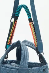 Cartera tote azul de nylon matelassé con diseño de corazones. Tiene manijas cortas y dos correas desmontables, una de cuero sintético y otra de tela trenzada multicolor. Cuenta con cierre superior y dos bolsillos internos.