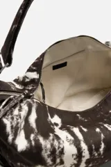 Bolso de hombro tipo bandolera, confeccionado en tejido de algodón con estampado abstracto en tonos blanco y negro. Cuenta con un compartimento principal con cierre de cremallera, un bolsillo interior con cremallera y asa de hombro ajustable.