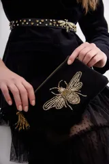 Bolso de mano tipo sobre rectangular de terciopelo negro con cierre dorado y flecos de mostacillas doradas. Presenta un bordado de abeja con canutillos dorados en el frente.