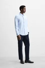 Camisa celeste a rayas verticales blancas, de corte recto, con cuello italiano abierto, manga larga con puños abotonados y cierre frontal con botones.