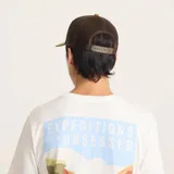 Gorro trucker marrón con paneles frontales rígidos, visera curva y paneles traseros de malla. Presenta un parche con el logo de Roark en el frente.