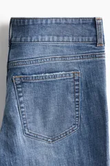 Jeans de denim azul medio con ligero efecto desgastado y roturas a la altura de las rodillas. Presenta un corte ajustado en muslos y acampanado desde la rodilla. El tiro es bajo, con pretina prolongada que se cierra con dos botones visibles. Incluye bolsillos delanteros tipo parche y bolsillos traseros.