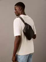 Riñonera Calvin Klein negra de cuero sintético texturizado, con cierre principal y logo CK grabado en el frente. La correa es ajustable y de color gris, con el nombre de la marca repetido.