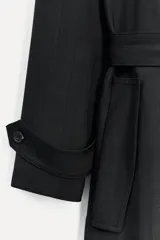 Trench negro oversize con cuello solapa, manga larga con trabilla y botón, bolsillos delanteros, cinturón ajustable con lazadas, abertura en la espalda y cierre frontal con botones ocultos por solapa.