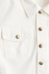 Mono corto color blanco de denim rígido, con cuello camisero, cierre abotonado, mangas cortas, bolsillos superiores con botón y cinturón extraíble.
