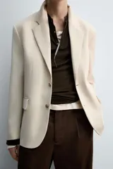Blazer color beige confeccionado en tejido con lana, de corte relajado. Tiene cuello con solapas de muesca, manga larga con puños abotonados, bolsillo de vivo en el pecho, bolsillos de solapa en la cadera, bolsillo interior, bajo con abertura en el centro de la espalda y cierre frontal con botones.