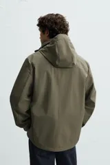 Parka verde militar de tejido técnico ligero resistente al viento, con cuello alto, capucha y manga larga con puños ajustables. Tiene bolsillos de plastrón con solapa en la cadera, bolsillo interior y bajo ajustable con elásticos. El cierre frontal es con cremallera oculta por solapa con botones a presión.
