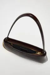 Bolso de hombro alargado color marrón oscuro con detalle metálico dorado en un lateral, asa corta y cierre de cremallera.