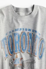 Remera gris de corte amplio con estampado de casco de fútbol americano y texto en azul y naranja.