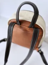 Mochila de cuero color tostado, con base y espalda en negro y tapa superior en blanco. Tiene un bolsillo frontal con cierre y un pequeño aplique de cuero con el logo de la marca.