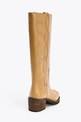 Bota alta de cuero color camel, con caña recta y punta redondeada. Presenta pespuntes decorativos verticales en la parte delantera y un tacón ancho de madera.