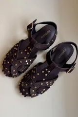 Sandalia tipo guillermina confeccionada en cuero nobuk color chocolate con tachas doradas y hebilla lateral.