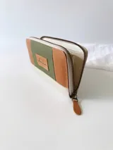 Billetera grande con cierre metálico, confeccionada en cuero con diseño de bloques de color: verde oliva texturizado, marrón claro liso y blanco/crema texturizado. Posee un monedero interno y un tirador de cuero marrón claro.