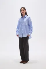 Camisa oversize de poliéster, con cuello camisero y cierre frontal de botones. Presenta un diseño de rayas verticales azules y blancas, con bloques de rayas horizontales en los puños y el ruedo inferior.
