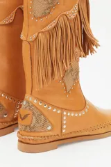 Bota alta color cuero con flecos, apliques de cuero con estampado reptil, tachas plateadas y costuras a la vista.