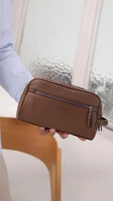 Neceser unisex marrón de símil cuero, con doble compartimento superior y bolsillo frontal con cierre. Asa lateral para fácil transporte.