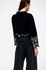 Blusa cropped de algodón color negro con bordado de flores blancas, mangas largas abullonadas, escote en V con volados y cierre frontal con lazos.