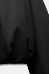Cazadora corta negra con cuello solapa y manga larga. Presenta detalle de acabados con pliegues y cierre frontal con cremallera.