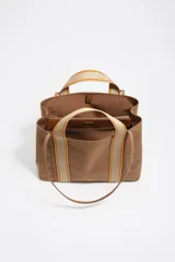 Bolso shopper grande de estructura blanda, color marrón, confeccionado en nylon con detalles de piel vacuna. Presenta doble asa de mano con cinta de tejido beige y naranja, y un logo metálico de Bimba y Lola en el frente.