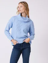 Buzo de punto grueso color beige, con cuello volcado y textura en relieve. Tiene mangas largas de corte hombro caído y puños anchos.