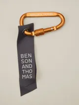 Llavero con mosquetón metálico color cobre y cinta de tela gris con el nombre de la marca impreso en blanco.