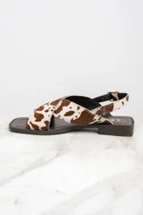 Sandalias bajas con tiras cruzadas de cuero con estampado de vaca en tonos marrón y blanco. Tira trasera con hebilla dorada y suela plana color marrón.