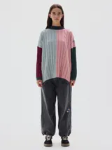 Buzo tejido colorblock con cuello subido tipo polera, diseño oversized y largo con pequeños cortes laterales. Presenta bloques de color rosa, celeste, bordó y verde oscuro, con cuello, puños y dobladillo en gris oscuro.
