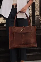 Bolso tipo tote de cuero color marrón con detalles de costuras trenzadas en los bordes y bolsillo externo. Tiene cierre imantado interno y broche con pasador de guampa en el exterior.
