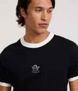 Remera de algodón negra, estilo ringer, con cuello y ribetes de manga en color blanco contrastante. Presenta un pequeño bordado de un ángel en el centro del pecho.