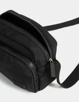 Bandolera negra de tela deportiva con bolsillo frontal con cierre, correa regulable y tag de goma.